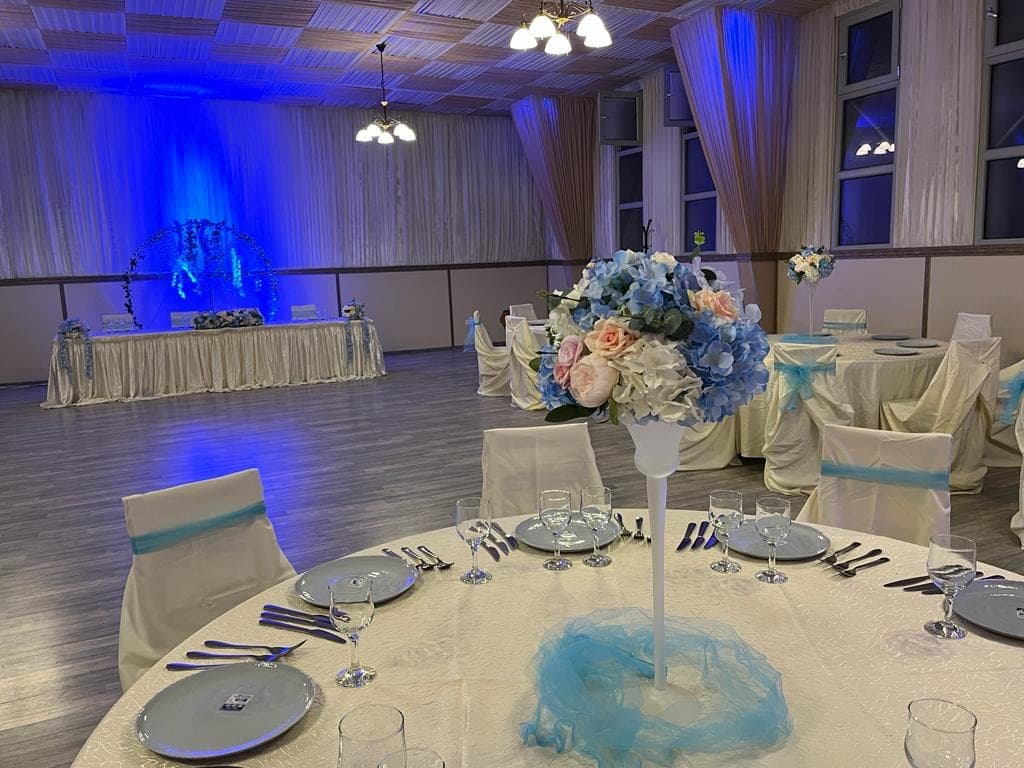 Mese rotunde aranjate pentru eveniment, cu aranjamente florale înalte și husele scaunelor legate cu organza bleu